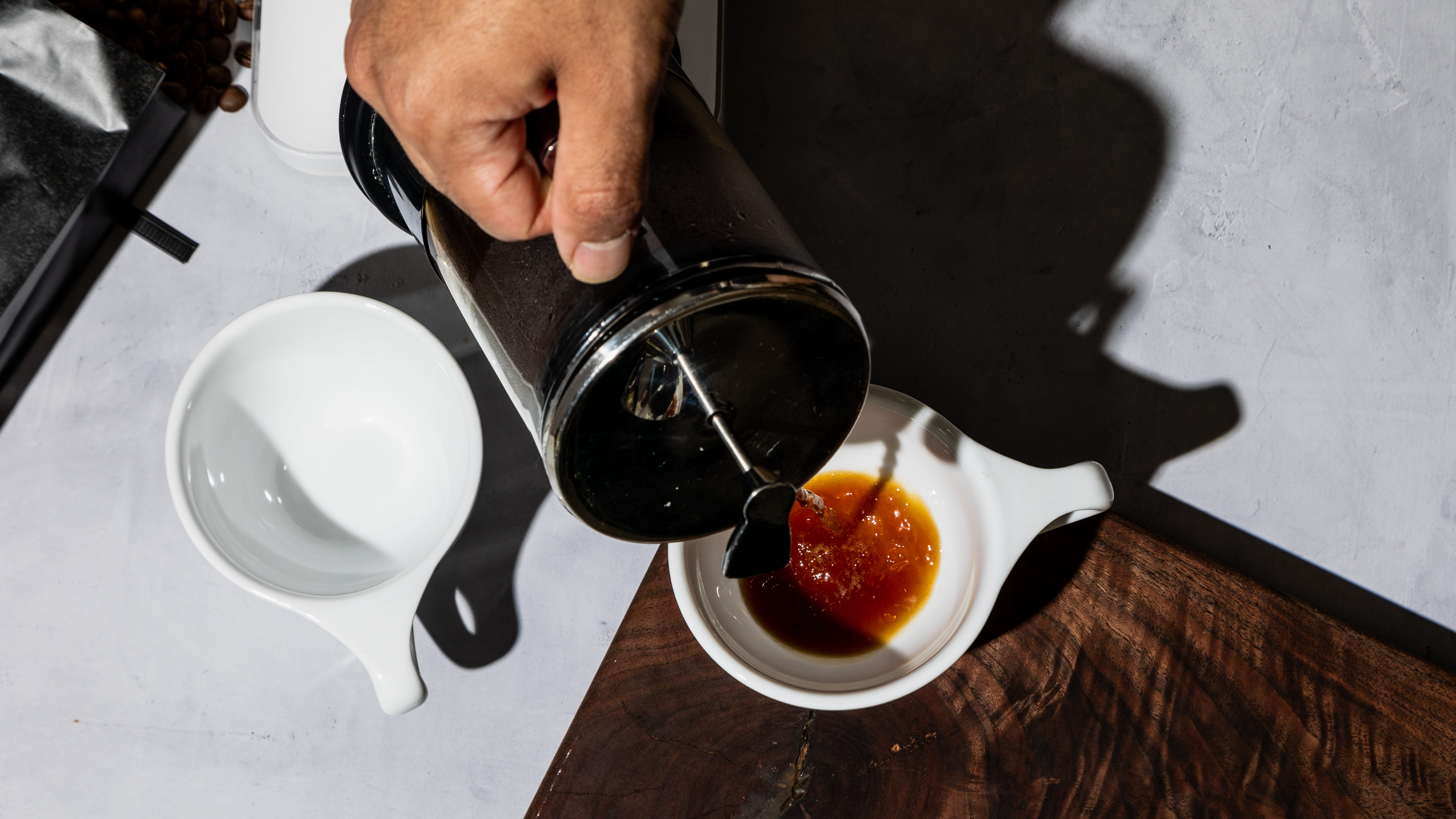 Person pouring coffee from a French press into a white cup on a wooden surface.